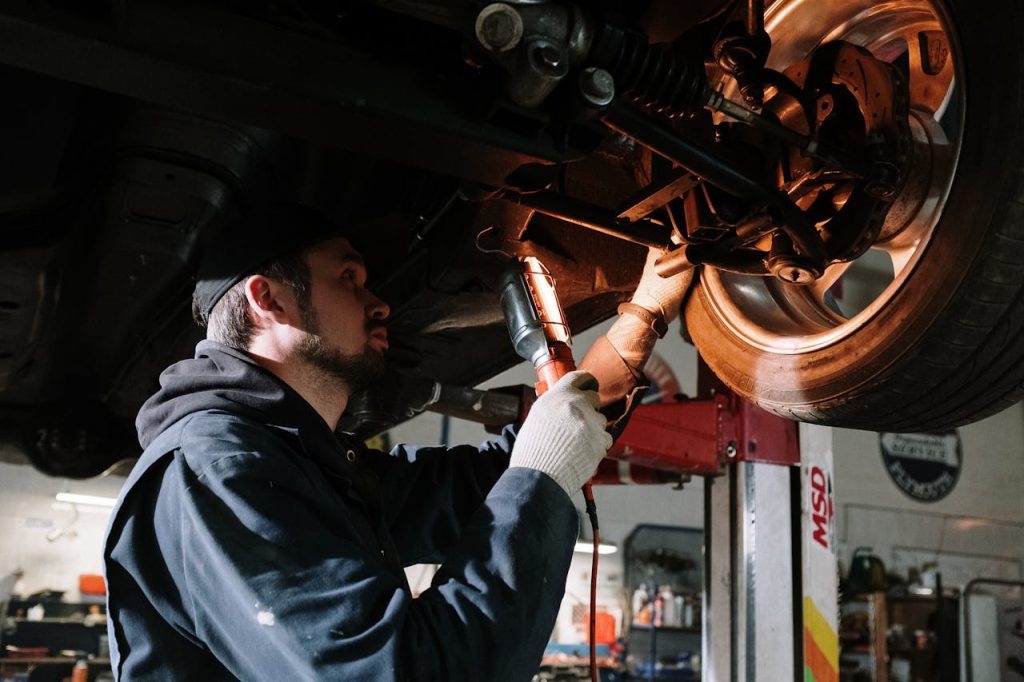 man-in-blue-dress-shirt-holding-red-and-black-power-tool-4489749 Mechanic examining cars undercarriage at a garage, focusing on vehicle maintenance.